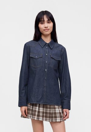 Junge Frau mit glatten dunklen Haaren trägt ein langärmeliges, dunkles Jeanshemd und einen plissierten braun-weiß karierten Rock.