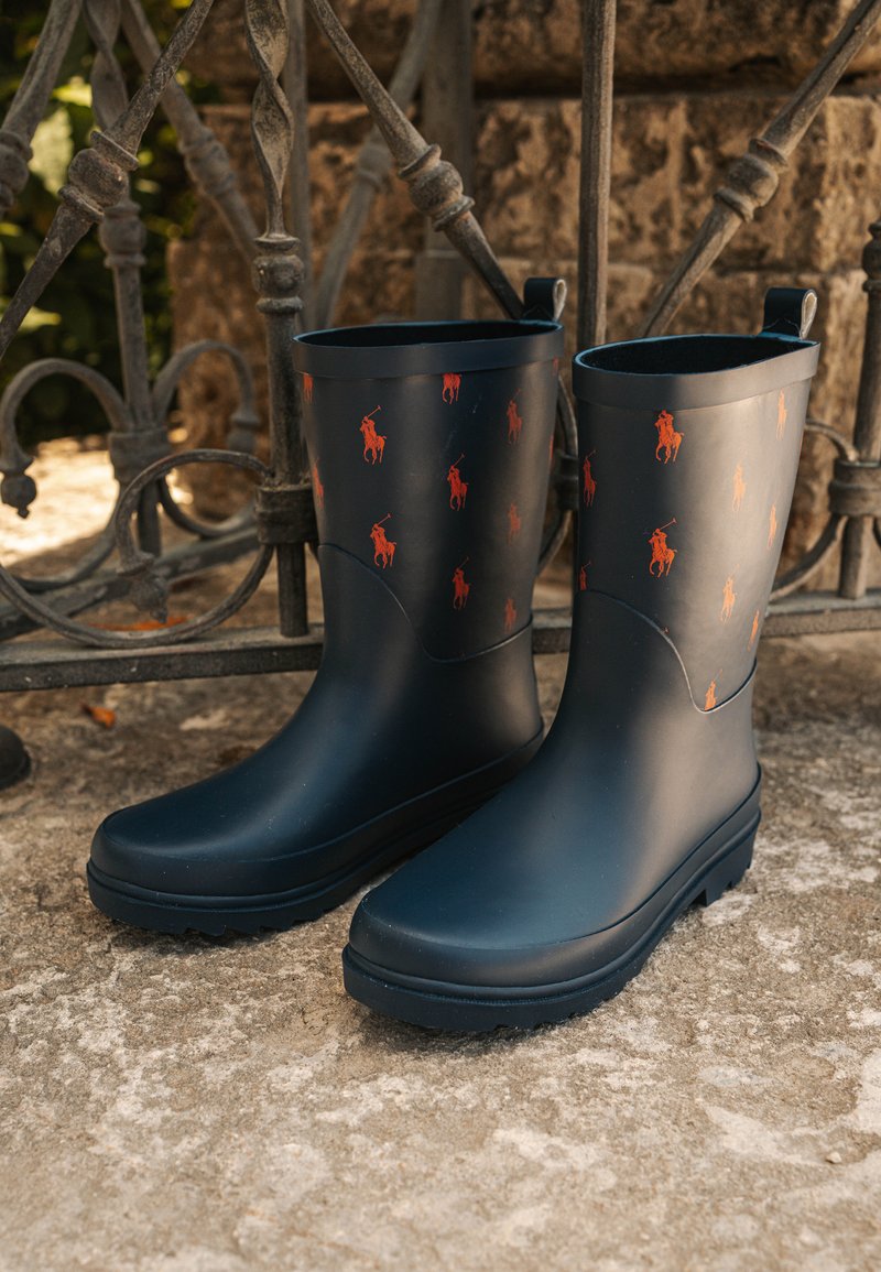 Bottes de pluie noires en caoutchouc avec des motifs de logo de cheval orange, semelles plates et tirants en boucle sur les côtés. Texture de surface lisse et bout arrondi.