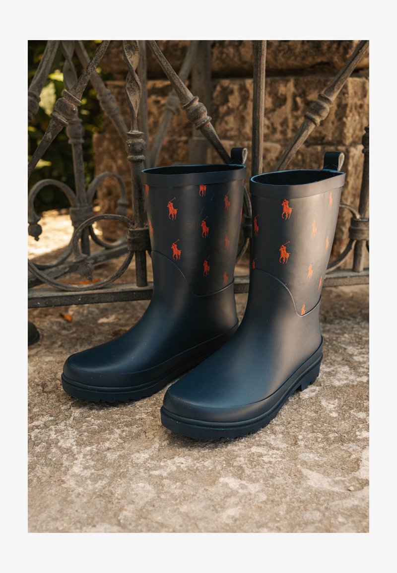 Bottes de pluie noires en caoutchouc avec des motifs de logo de cheval orange, semelles plates et tirants en boucle sur les côtés. Texture de surface lisse et bout arrondi.