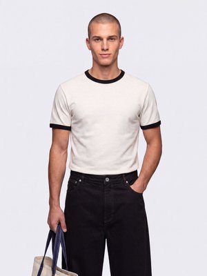 Jeune homme aux cheveux courts portant un t-shirt blanc avec des bordures noires et un pantalon noir, tenant un sac beige avec des poignées bleues.
