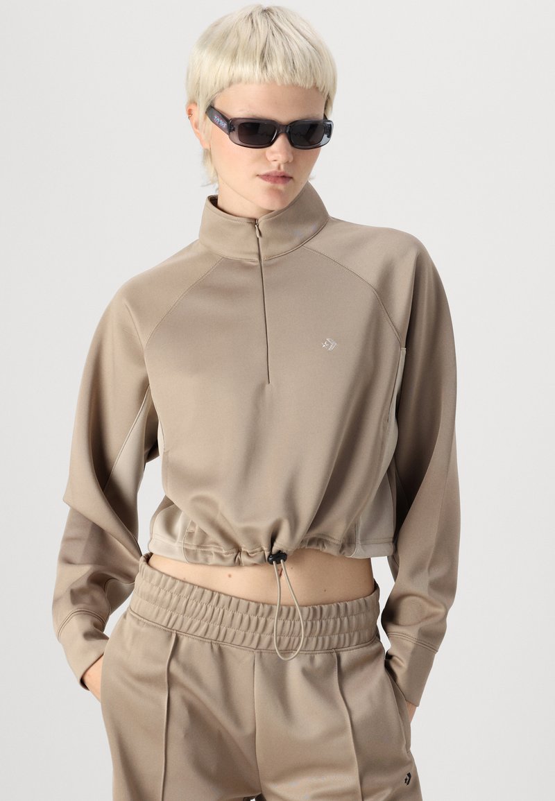 Felpa cropped beige con colletto alto, chiusura a zip e orlo con cordino. Presenta maniche lunghe e un logo sul petto.