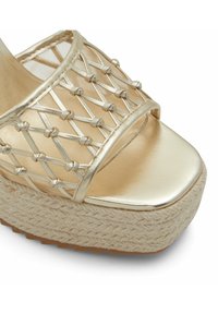 Zlaté platformové sandály s tkaným slámovým základem. Metalické pásky mají křížový vzor. Hladký, lesklý svršek s otevřenou špičkou.