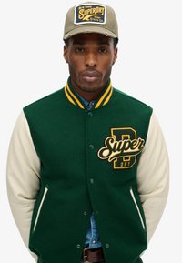 Chaqueta varsity de lana verde con mangas crema, con cuello de rayas amarillas y logo bordado "Super Dry" en el pecho. Botones marrones visibles.