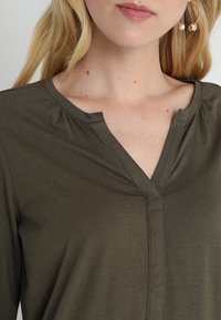 Blonde vrouw die een donkergroene V-hals top en grote gouden oorringen draagt, met een blootgestelde nek en bovenste deel van de borst, voorzien van lichte huidvlekken.