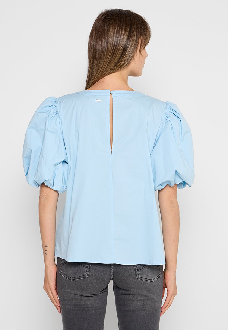 Helle blaue Bluse mit Puffärmeln, aus Baumwollmaterial, verfügt über eine Schlüsselöffnung auf der Rückseite und ein kleines Etikett am Ausschnitt.