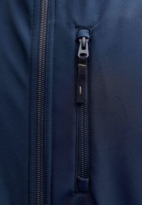 Nærbilde av marineblå jakke stoff med to svarte glidelåser, en vertikal og en horisontal glidelås til lomme med en stofftrekk.