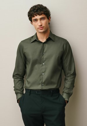 Young man with curly dark hair wearing olive green button-up shirt and dark pants, standing with hands in pockets against plain wall.