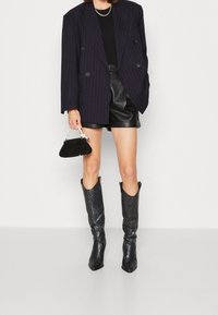 Musta raitakuviollinen kaksirivinen bleiseri, istuvat mustat nahkashortsit, teksturoidut mustat polven korkeudella olevat saappaat, joissa on terävä kärki, ja pieni clutch-laukku.