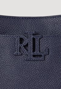 Jūras spēku teksturēta āda ar iepresēto "RL" logotipu. Tuvplāns, kurā redzams graudu raksts un šuvju detaļa izstrādājuma augšējā malā.