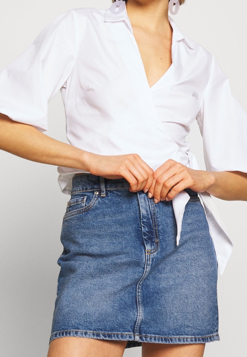 Femme portant un chemisier blanc portefeuille à manches bouffantes et une minijupe en jean bleue, ajustant la jupe à la taille.