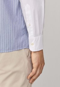 Mano maschile accanto alla tasca dei pantaloni beige, indossa una camicia a righe verticali blu con maniche lunghe a fascia bianca.