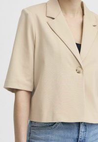 Beiger Kurzarm-Blazer mit einem Knopf, getragen über einem schwarzen Top und kombiniert mit blauen Jeans.