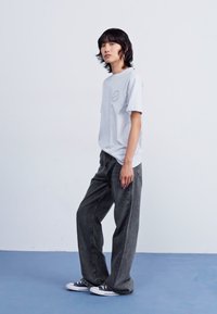 Maglietta in cotone grigia chiara con un logo circolare sul petto, abbinata a jeans grigi scuri a gamba larga e sneakers in bianco e nero.