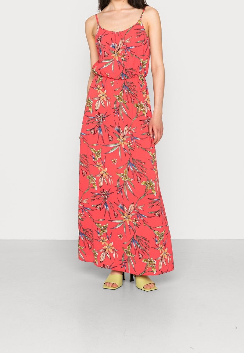 Femme portant une robe maxi corail à fines bretelles et motif floral coloré, associée à des talons ouverts vert clair.