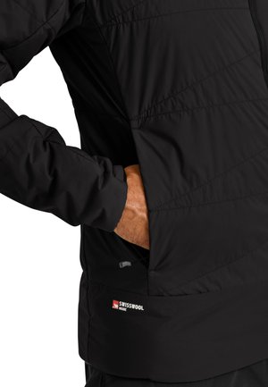 Hand in pocket of black quilted jacket labeled "Swisswool Inside" on lower side, showing jacket's texture and stitching details.