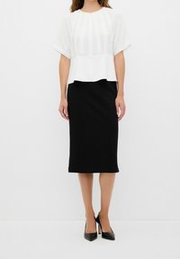 Femme portant un chemisier blanc à manches courtes, une jupe crayon noire longueur genou et des escarpins noirs à bout pointu, debout devant un fond uni.