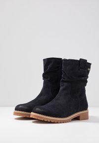 Botas negras de ante hasta el tobillo con un caña relajada, suela de goma beige y detalle de botón lateral. Presentan un acabado texturizado y diseño de punta redondeada.