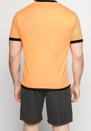 Homme portant un t-shirt de sport orange à manches courtes avec bordure noire et un short gris foncé, vu de dos sur un fond gris uni.