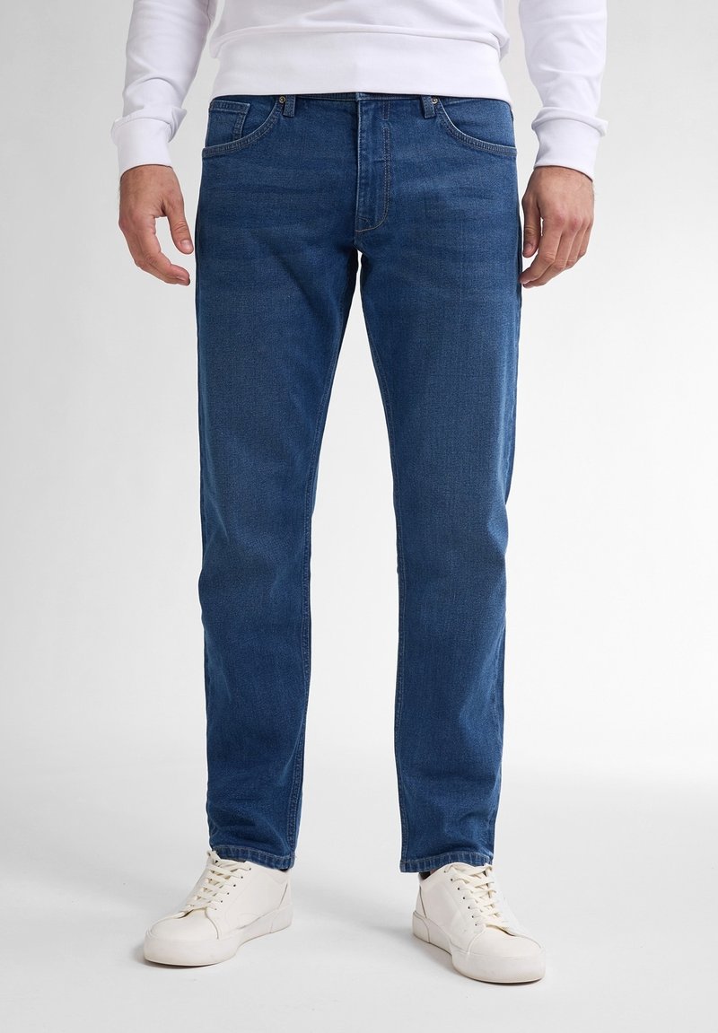 Jeans in denim blu con vestibilità dritta. Presentano cinque tasche, cuciture sottili e una chiusura lampo classica. Indossati con sneakers bianche.