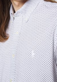 Detailní záběr na osobu v bílé košili na knoflíky s malým tmavým geometrickým vzorem a košilovým límcem.