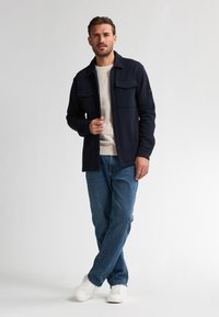 Marineblauwe button-up jas met twee borstzakken, gedragen over een lichtbeige trui, gekoppeld aan blauwe jeans en witte sneakers.