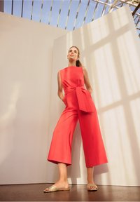 Tutina rossa con corpino aderente, ampie gambe culotte e cintura legata in vita. Modella che indossa sandali piatti con dettagli dorati, in piedi su uno sfondo chiaro.