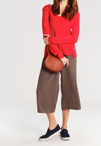 Czerwony sweter z długim rękawem, brązowe spodnie z szerokimi nogawkami, granatowe buty na slip-on oraz brązowa torebka o zaokrąglonym kształcie. Prosty design, gładka tekstura i neutralne kolory.