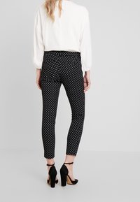 Svarta mönstrade byxor med små vita diamantformer, slim fit, ankellängd, tillsammans med svarta högklackade skor. Vit topp synlig bakåt.