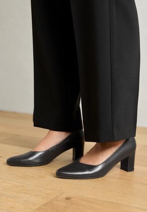 Sorte læder højhælte sko med firkantet tå og blokhæl, der har en glat tekstur og minimalistisk design, båret med sorte bukser.