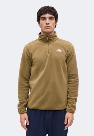 Ung mand iført en olivengrøn quarter-zip fleece fra The North Face og mørkeblå bukser, stående med fronten mod kameraet mod en ensfarvet baggrund.