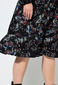 Musta mekko, jossa on värikäs perhoskuvio, röyhelöhelma ja teksturoitu kangas. Käsi lepää kankaan päällä lähellä helman reunaa.