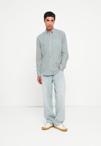 Chemise rayée boutonnée en bleu clair et blanc, associée à un jean en denim clair et des baskets blanches avec une semelle beige. Coupe décontractée, manches longues.