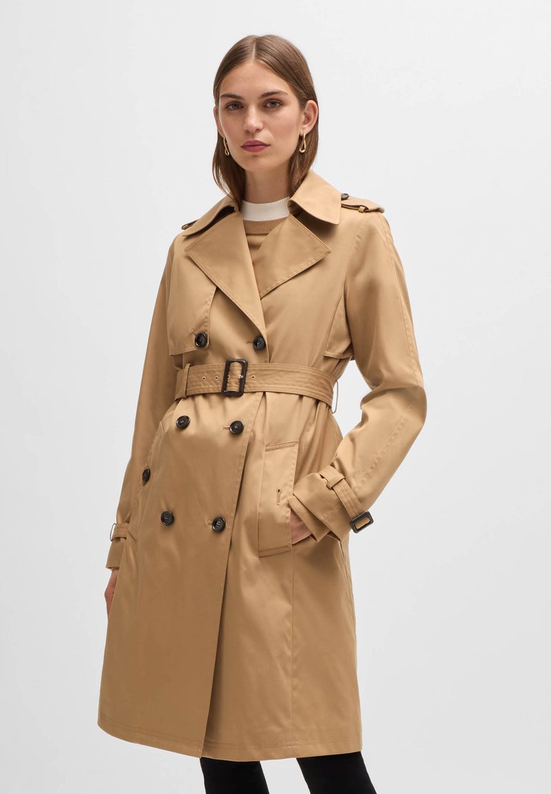 Beige Trenchcoat aus glattem Stoff, mit einem doppelt geknöpften Front, breiten Revers und einem Gürtel in der Taille. Schwarze Knöpfe und seitliche Taschen.