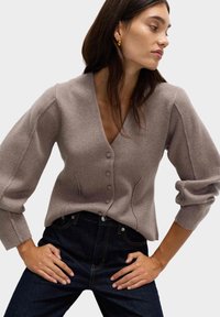 Helle beige gestrickte Pullover mit V-Ausschnitt, Knöpfen vorne und lockeren Ärmeln. Kombiniert mit dunkelblauen Jeans.