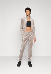 Fløyelstreningsdrakt i lys beige med en cropped jakke og bukser med elastisk midje, med ribbestrikkede mansjetter og snøring. Svarte joggesko.