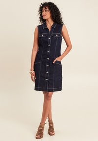 Robe en denim sans manches de couleur bleu foncé, avec poches poitrine, quincaillerie en métal argenté, boutons à l'avant et couture blanche. Longueur mi-cuisse.