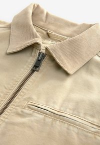 Close-up of beige jacket featuring a corduroy collar, front zipper closure, and horizontal chest pocket.