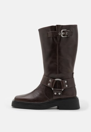 Barna bőr, térd fölé érő csizma, lekerekített orral, széles pánttal, amely fémhardverrel díszített, és egy erős fekete talppal. Simaságával kiemelkedő textúra.