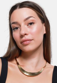Collana in metallo dorato, curvata, con una finitura lucida. Presenta una chiusura in metallo, indossata sulla pelle nuda contro una maglietta scura.