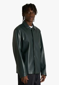 Giacca in pelle verde scuro con collo a risvolto, maniche lunghe e chiusura frontale con zip. Texture liscia con finitura leggermente lucida.