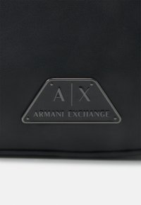 Czarna torba z imitacji skóry z trójkątnym srebrnym metalowym logo z wygrawerowanym napisem „A/X” i „Armani Exchange”. Gładka faktura.