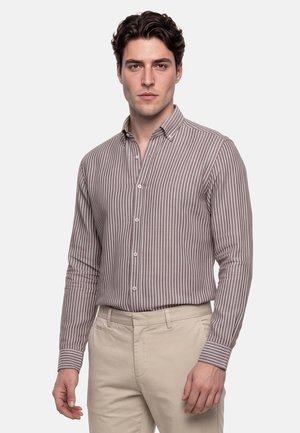 Mand iført en brun og hvid vertikalt stribet skjorte med knapper og beige bukser, stående mod en ensfarvet lys baggrund.