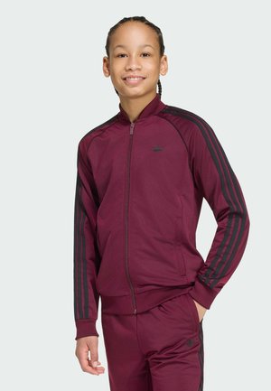 Junge Person mit geflochtenen Haaren, die einen maroonfarbenen Adidas-Trainingsanzug mit schwarzen Streifen trägt, lächelt und mit einer Hand in der Tasche steht.