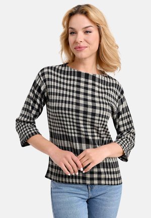 Schwarz-weißer karierter Oberteil mit 3/4-Ärmeln, rundem Halsausschnitt und weichem Material. Kombiniert mit hellblauen Jeans, die Hände sind lässig positioniert.
