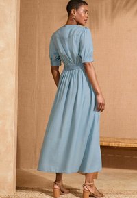 Light blue, knee-length dress with puff sleeves, a fitted waist, and flowing skirt. Worn with tan strappy heels on a textured surface.