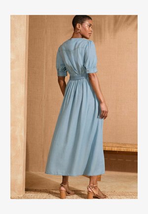 Abito azzurro chiaro, fino al ginocchio, con maniche a palloncino, vita aderente e gonna fluida. Indossato con tacchi beige con cinturini su una superficie texturizzata.