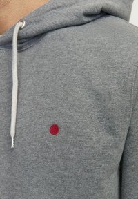 Felpa grigia con cappuccio e cordini, caratterizzata da un piccolo punto rosso ricamato sul petto. Tessuto morbido e design casual.