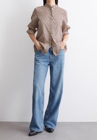 Brązowa bluzka w kremowy wzór z kołnierzykiem mandarynki i bufiastymi rękawami, zestawiona z jasnoniebieskimi spodniami o szerokich nogawkach i czarnymi, spiczastymi butami.