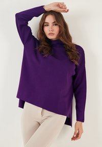 Lila Strickpullover mit hohem Kragen, lässiger Passform und Seitenschlitzen. Getragen mit beigen Hosen, die lange Ärmel und eine glatte Textur aufweisen.