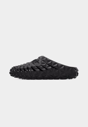 Zapato negro de tipo slip-on con patrón geométrico de triángulos y ranuras de ventilación en ángulo en el lateral, mostrado en vista de perfil.